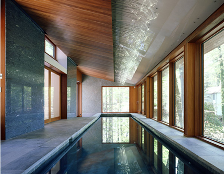 Photo: Catherine Tighe.  Due to the slope of the site, the swimmer is 12’ above grade at the deep end of the pool near the window.  The pool finish, applied by hand, is a tinted plaster in a bluish-grey color achieved by mixing several standard dry pigments.  The room is heated by the pool water and the radiant heat in the concrete floor slab.  The floor and pool coping is green quartzite tile; the walls are a combination of the Brazilian Azul du Mar marble and the green Costa Esmerelda granite.  The wood frame of the Duratherm sliding and fixed doors and windows is in a clear finish mahogany; the tongue-and-groove ceiling is made from sapelle, as are the wood panels above the east-facing openings.  The glass ceiling panels are laminated with a translucent inner layer, minimally gapped for air flow, and held in place to a Uni-Strut steel system above with stainless steel Tri-Pyramid standoffs.  Leveling of the glass panels was challenging and was made possible by the adjustable anchors.  At the ceiling plane change, 520 stainless steel rods threaded with fiber-optic strands light the space.  The dots of light reflect off the ceiling glass, the wall glazing and the water, particularly as the outside light level dims.